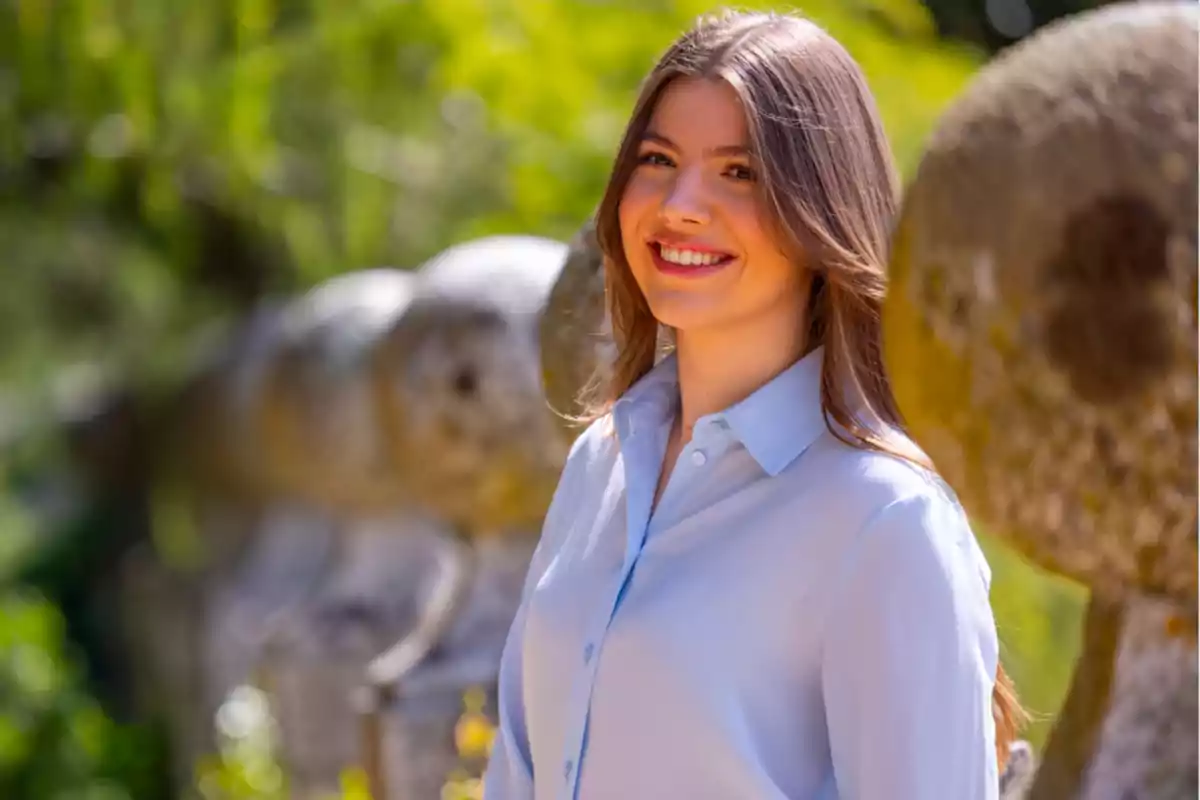 Una mujer joven con cabello castaño y camisa azul claro sonríe al aire libre con un fondo de vegetación y piedras redondeadas.