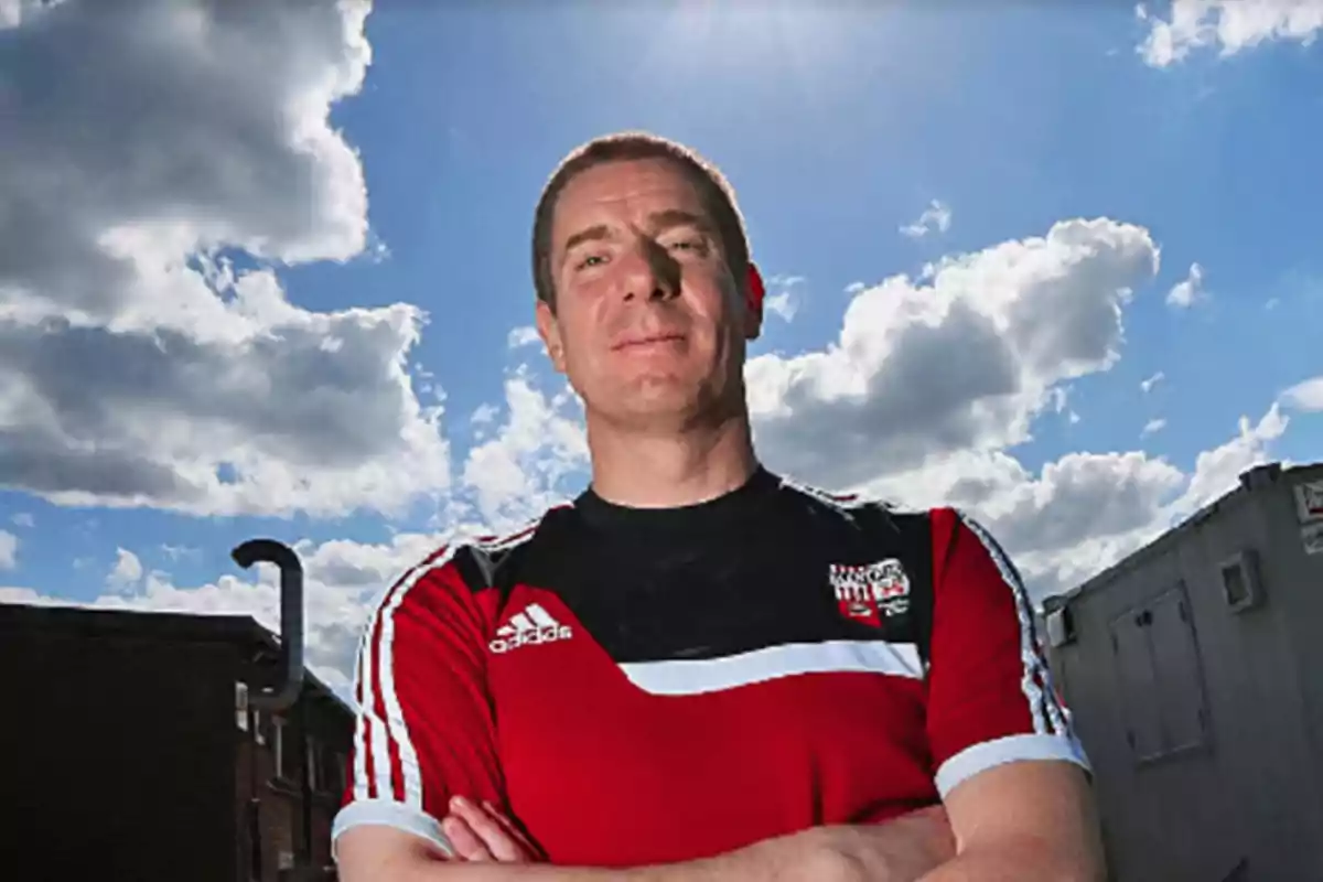 Un hombre con camiseta deportiva roja y negra está de pie al aire libre con los brazos cruzados bajo un cielo parcialmente nublado. Un hombre con camiseta deportiva roja y negra está de pie al aire libre con los brazos cruzados bajo un cielo parcialmente nublado.