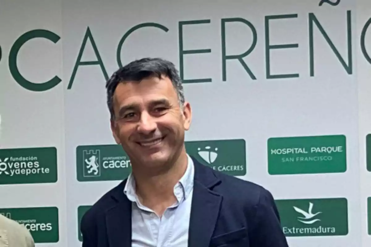 Un hombre sonriendo frente a un fondo con logotipos y texto relacionado con Cáceres y Extremadura. Un hombre sonriendo frente a un fondo con logotipos y texto relacionado con Cáceres y Extremadura.