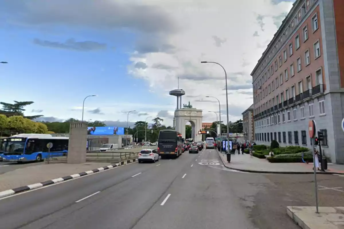 Una calle con tráfico y un arco monumental al fondo, junto a un edificio grande y un cielo parcialmente nublado. Una calle con tráfico y un arco monumental al fondo, junto a un edificio grande y un cielo parcialmente nublado.