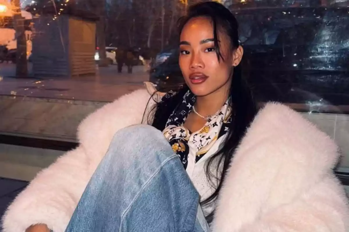 Mujer con abrigo blanco y jeans sentada frente a una ventana en un entorno urbano nocturno.