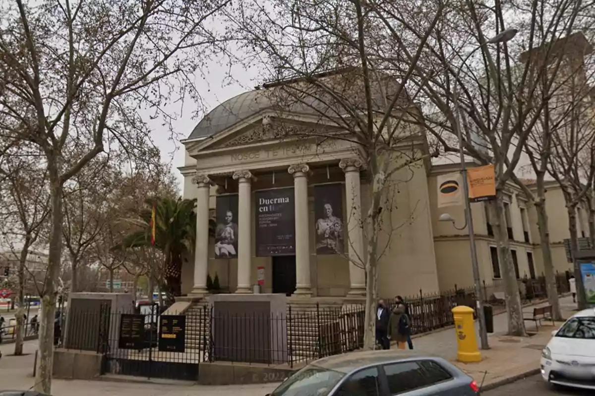 Edificio histórico con columnas y cúpula rodeado de árboles y personas caminando por la acera.