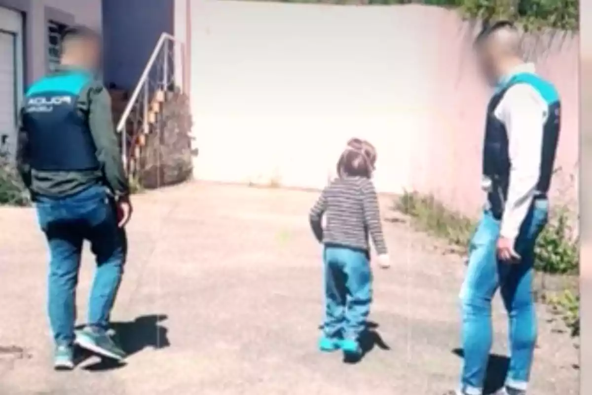 Dos adultos con chalecos acompañan a un niño mientras caminan por un camino pavimentado cerca de un edificio. Dos adultos con chalecos acompañan a un niño mientras caminan por un camino pavimentado cerca de un edificio.