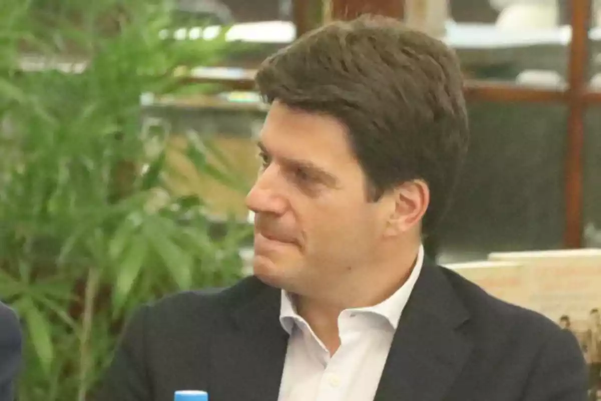 Un hombre de cabello oscuro y traje oscuro está sentado en un entorno interior con plantas al fondo. Un hombre de cabello oscuro y traje oscuro está sentado en un entorno interior con plantas al fondo.