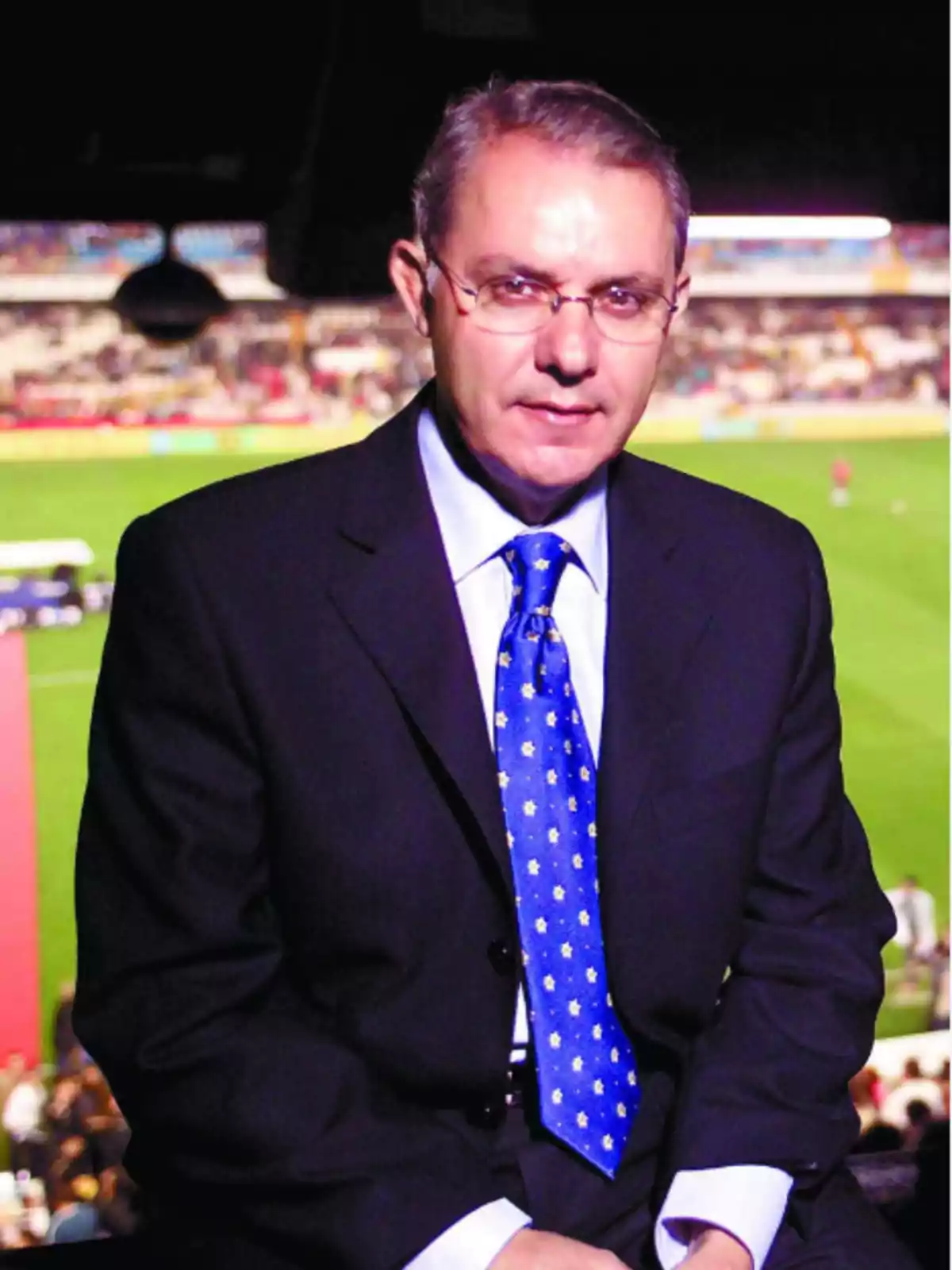 Un hombre con traje oscuro y corbata azul está sentado frente a un estadio de fútbol iluminado. Un hombre con traje oscuro y corbata azul está sentado frente a un estadio de fútbol iluminado.