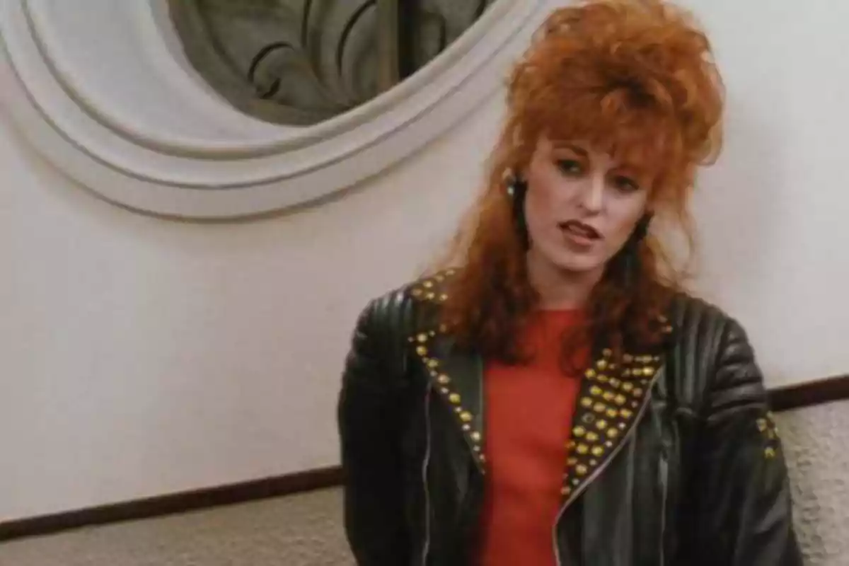 Una persona con cabello rojo y voluminoso lleva una chaqueta de cuero con tachuelas doradas y una camiseta roja, de pie frente a una pared blanca con un espejo decorativo. Una persona con cabello rojo y voluminoso lleva una chaqueta de cuero con tachuelas doradas y una camiseta roja, de pie frente a una pared blanca con un espejo decorativo.