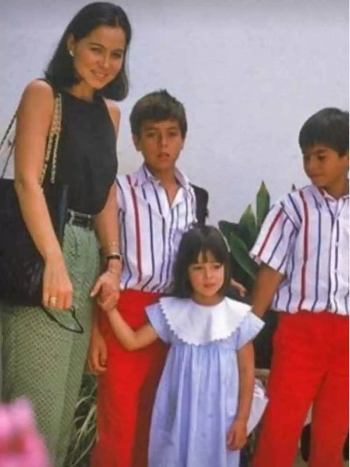 Una mujer sonriente sostiene la mano de una niña pequeña mientras dos niños están de pie junto a ellas, todos posando para la foto frente a un fondo blanco y plantas. Una mujer sonriente sostiene la mano de una niña pequeña mientras dos niños están de pie junto a ellas, todos posando para la foto frente a un fondo blanco y plantas.