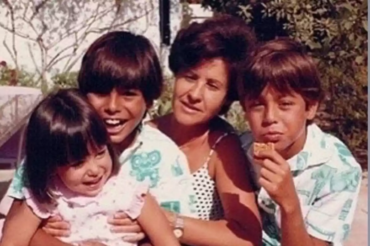 Una mujer sonriente rodeada de tres niños, dos de ellos abrazándola y uno comiendo una galleta, en un ambiente al aire libre. Una mujer sonriente rodeada de tres niños, dos de ellos abrazándola y uno comiendo una galleta, en un ambiente al aire libre.