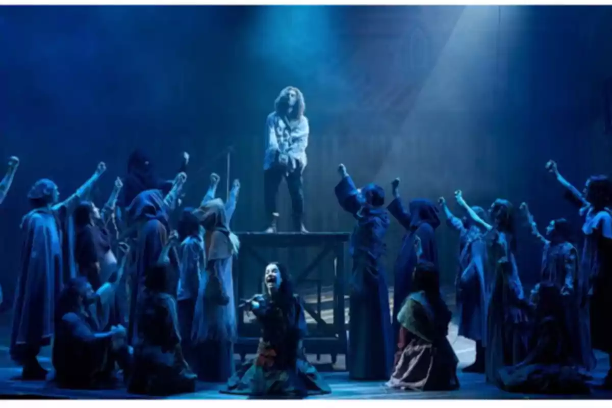 Un grupo de actores en una escena teatral dramática con iluminación azul y un personaje central elevado. Un grupo de actores en una escena teatral dramática con iluminación azul y un personaje central elevado.