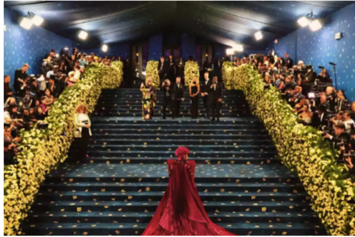 Una persona con un vestido rojo sube unas escaleras decoradas con flores amarillas, rodeada de fotógrafos y espectadores.