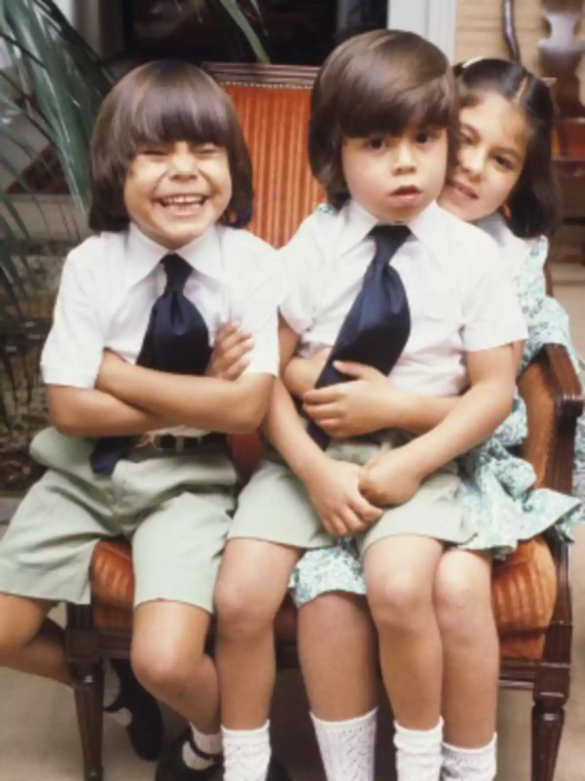 Tres niños sentados en una silla, dos de ellos con camisas blancas y corbatas oscuras, mientras una niña los abraza desde atrás, todos con expresiones alegres. Tres niños sentados en una silla, dos de ellos con camisas blancas y corbatas oscuras, mientras una niña los abraza desde atrás, todos con expresiones alegres.