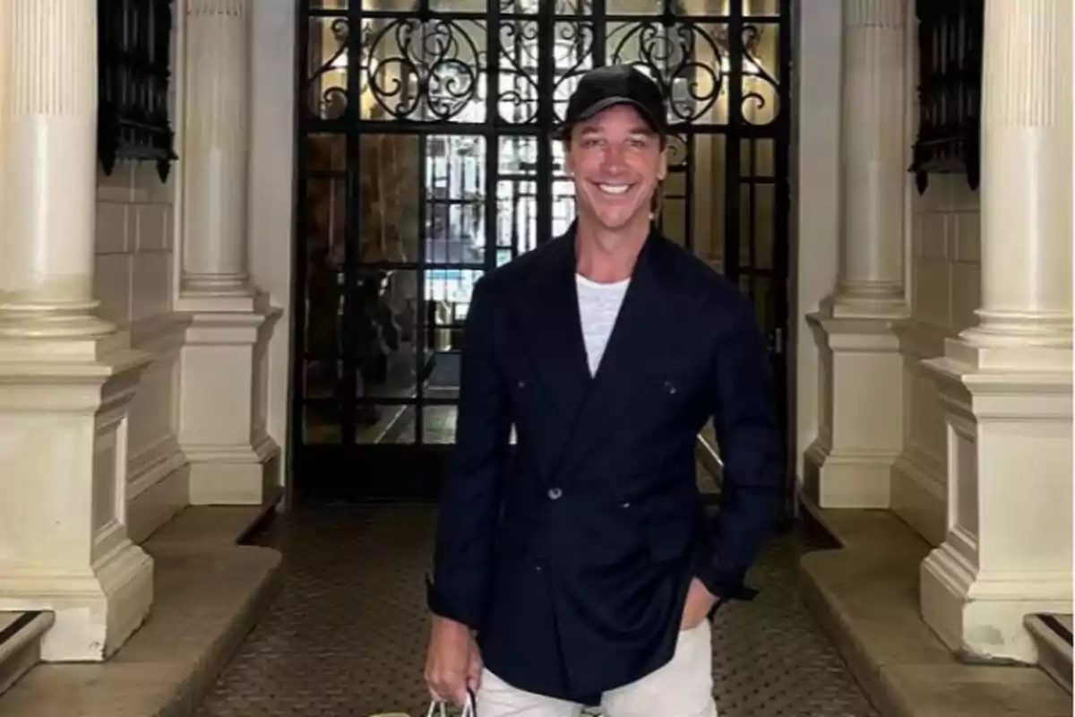 Un hombre sonriente con gorra y chaqueta oscura está de pie frente a una puerta de hierro forjado en un edificio con columnas. Un hombre sonriente con gorra y chaqueta oscura está de pie frente a una puerta de hierro forjado en un edificio con columnas.