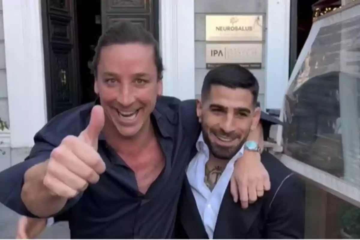 Dos hombres sonrientes posan juntos frente a un edificio, uno de ellos levanta el pulgar en señal de aprobación. Dos hombres sonrientes posan juntos frente a un edificio, uno de ellos levanta el pulgar en señal de aprobación.