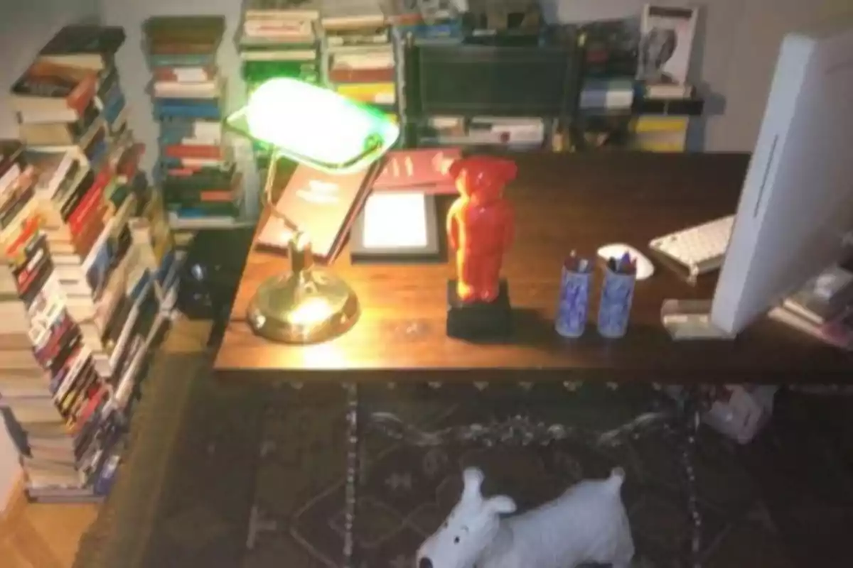 Una oficina con un escritorio de madera, una lámpara de escritorio verde, una figura decorativa roja, varios lápices en recipientes, una computadora y pilas de libros alrededor, además de una figura de un perro blanco en el suelo. Una oficina con un escritorio de madera, una lámpara de escritorio verde, una figura decorativa roja, varios lápices en recipientes, una computadora y pilas de libros alrededor, además de una figura de un perro blanco en el suelo.
