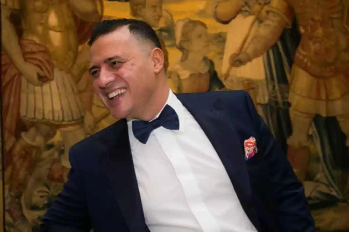 Un hombre sonriente con esmoquin y pajarita en un ambiente elegante con un tapiz de fondo.