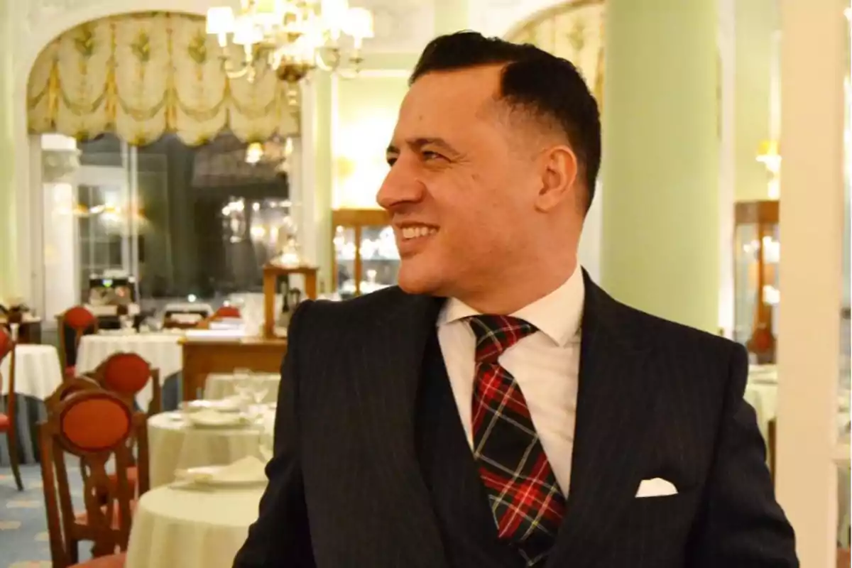 Un hombre sonriente con traje oscuro y corbata a cuadros está de pie en un elegante restaurante con mesas redondas y sillas rojas.