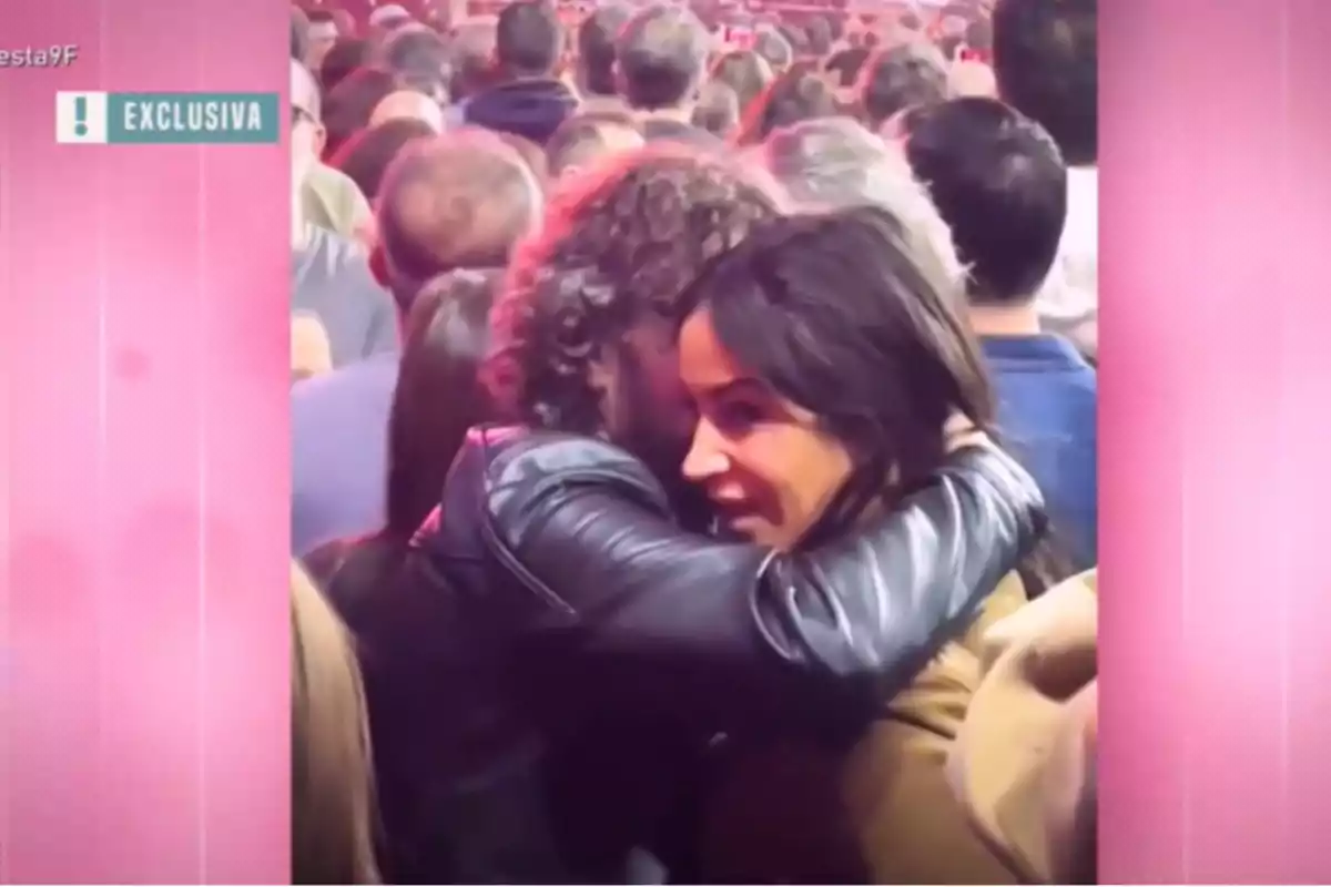Una pareja se abraza en medio de una multitud durante un evento, con un ambiente iluminado por luces rosadas.