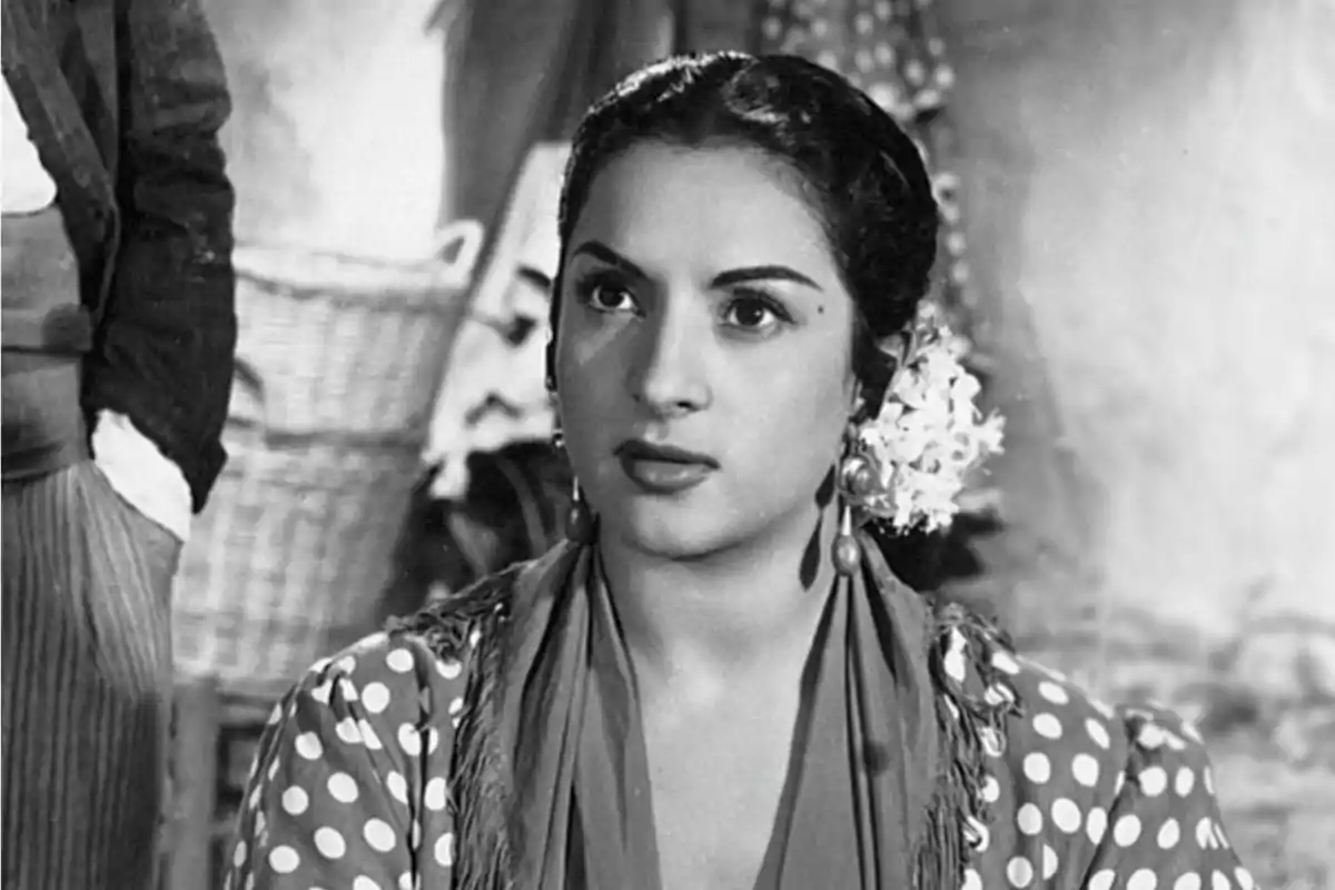 Mujer con peinado tradicional y flores en el cabello usando un vestido de lunares y pendientes grandes en una escena en blanco y negro.