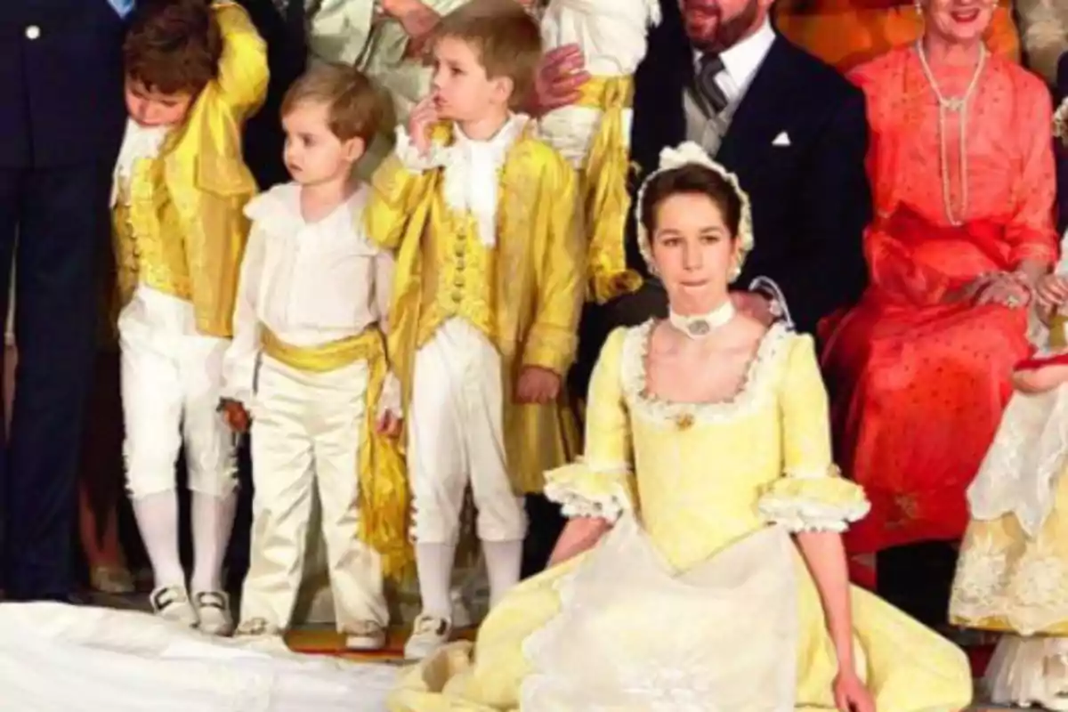 Un grupo de personas vestidas con trajes de época, incluyendo niños con chaquetas doradas y pantalones blancos, y una joven con un vestido amarillo claro, posan para una foto en un evento formal. Un grupo de personas vestidas con trajes de época, incluyendo niños con chaquetas doradas y pantalones blancos, y una joven con un vestido amarillo claro, posan para una foto en un evento formal.