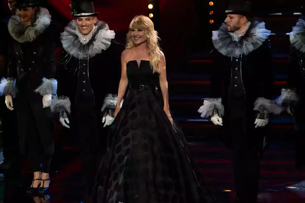 Un grupo de personas vestidas con trajes elegantes y accesorios teatrales en un escenario iluminado.