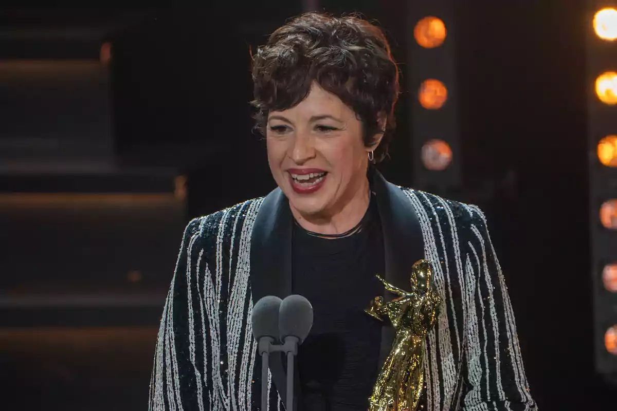 Una persona sonriente con cabello corto y rizado sostiene un trofeo dorado mientras habla frente a un micrófono en un escenario iluminado. Una persona sonriente con cabello corto y rizado sostiene un trofeo dorado mientras habla frente a un micrófono en un escenario iluminado.