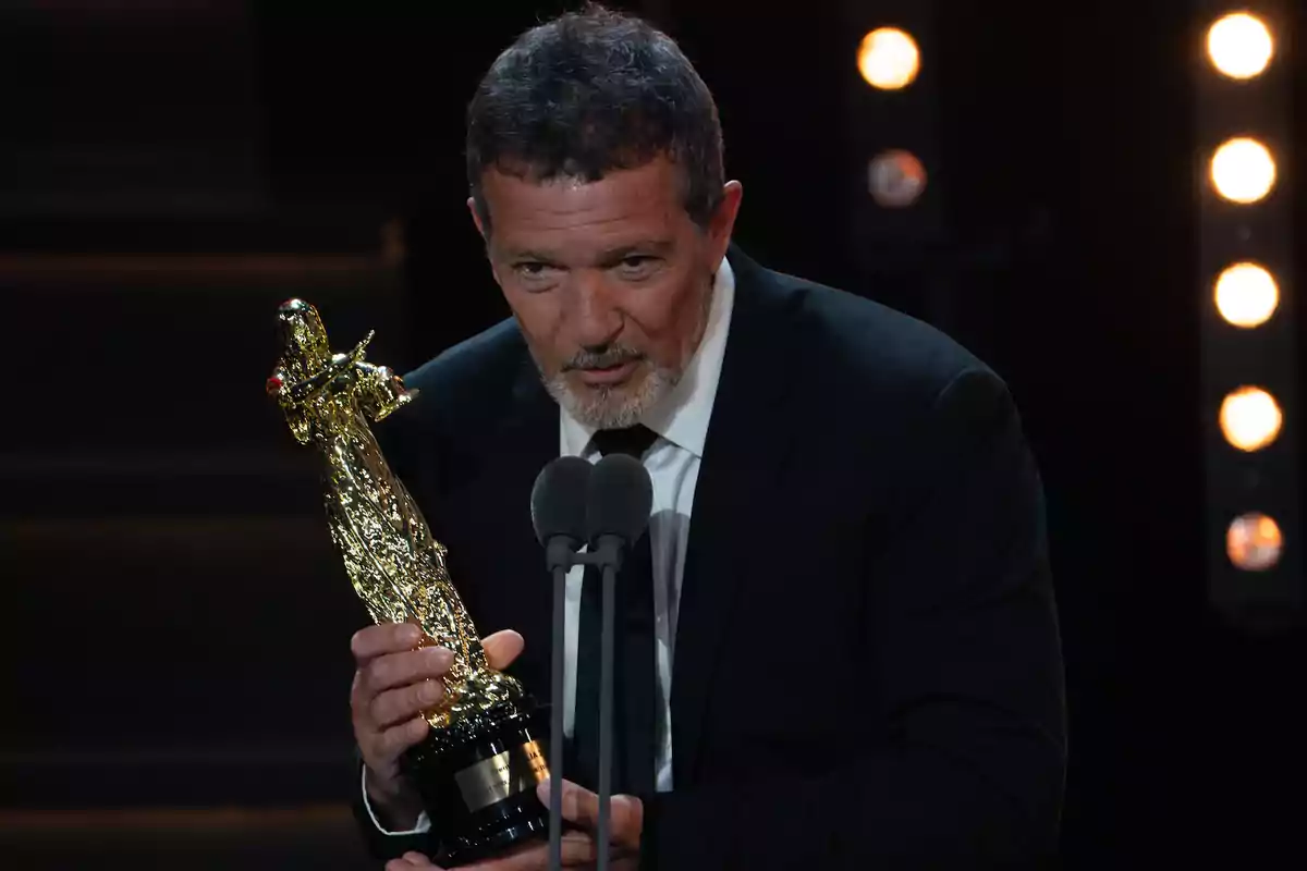 Un hombre con barba y traje oscuro sostiene un trofeo dorado mientras habla frente a un micrófono en un escenario iluminado. Un hombre con barba y traje oscuro sostiene un trofeo dorado mientras habla frente a un micrófono en un escenario iluminado.