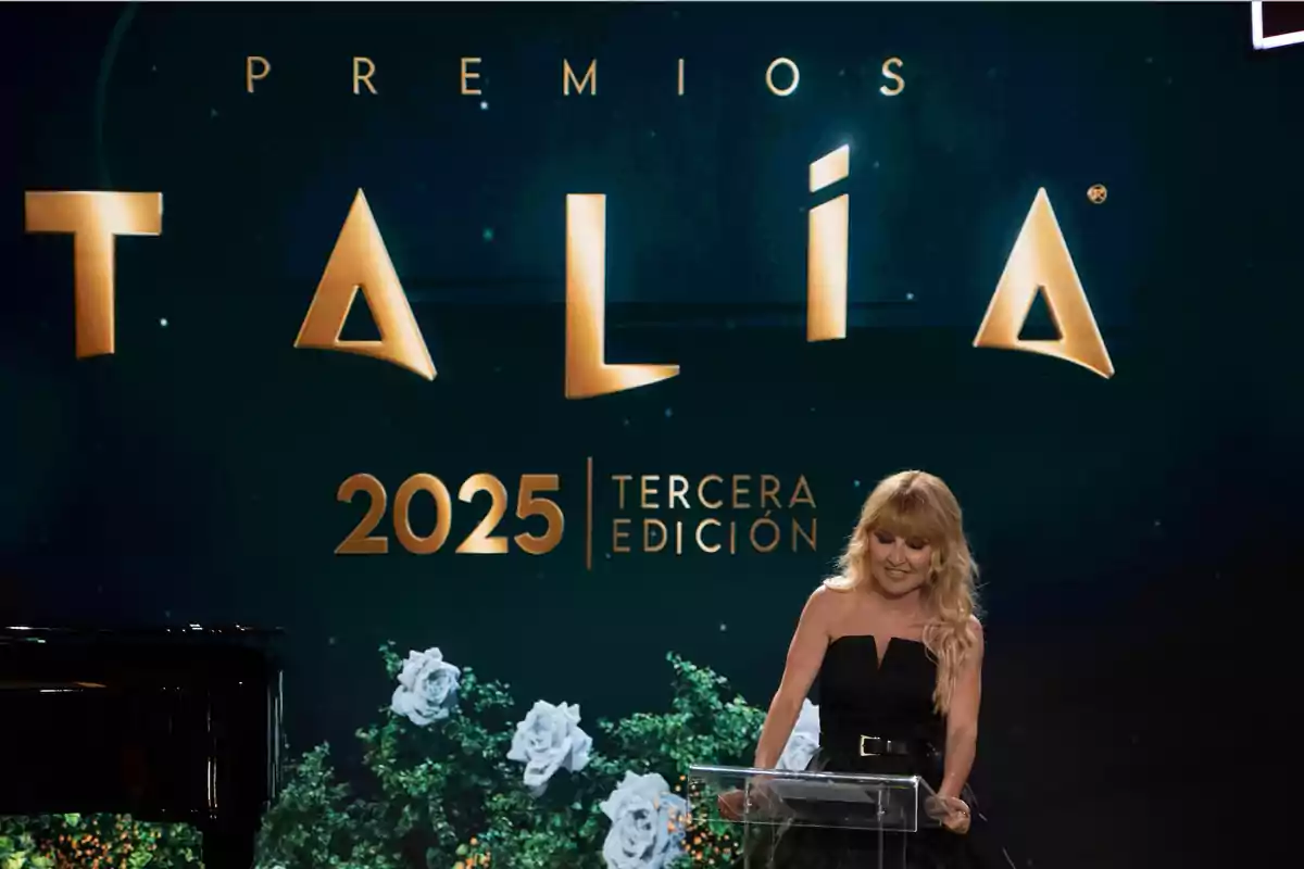 Una mujer de cabello rubio y vestido negro está de pie en un podio durante la ceremonia de los Premios Talia 2025 Tercera Edición con un fondo decorado con letras doradas y flores blancas. Una mujer de cabello rubio y vestido negro está de pie en un podio durante la ceremonia de los Premios Talia 2025 Tercera Edición con un fondo decorado con letras doradas y flores blancas.