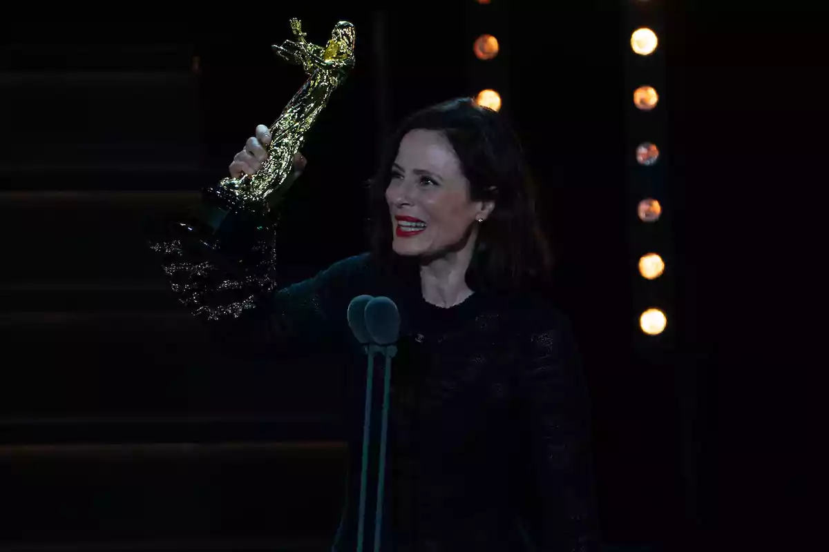 Una persona sonriente sostiene un trofeo dorado en un escenario iluminado. Una persona sonriente sostiene un trofeo dorado en un escenario iluminado.