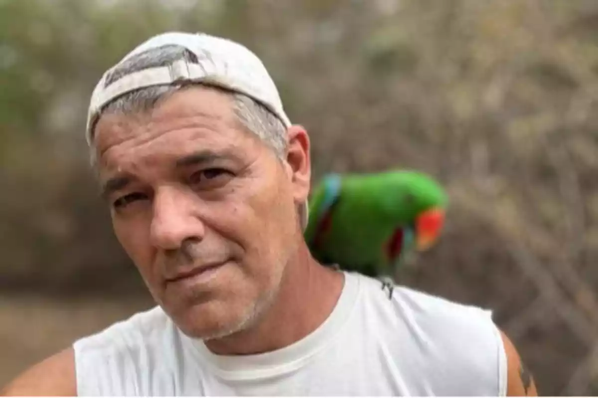 Un hombre con gorra blanca y camiseta sin mangas tiene un loro verde posado en su hombro.