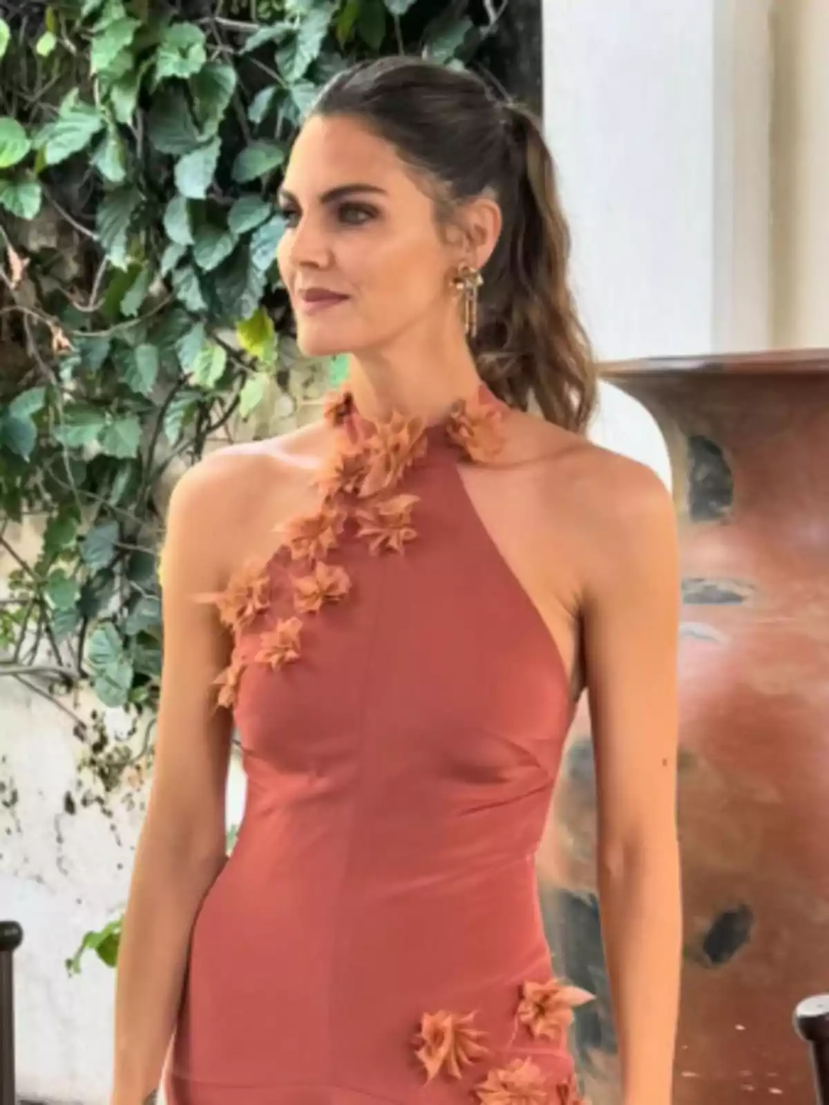 Una mujer con un vestido color terracota decorado con flores, de pie frente a un fondo de hojas verdes y una pared blanca. Una mujer con un vestido color terracota decorado con flores, de pie frente a un fondo de hojas verdes y una pared blanca.