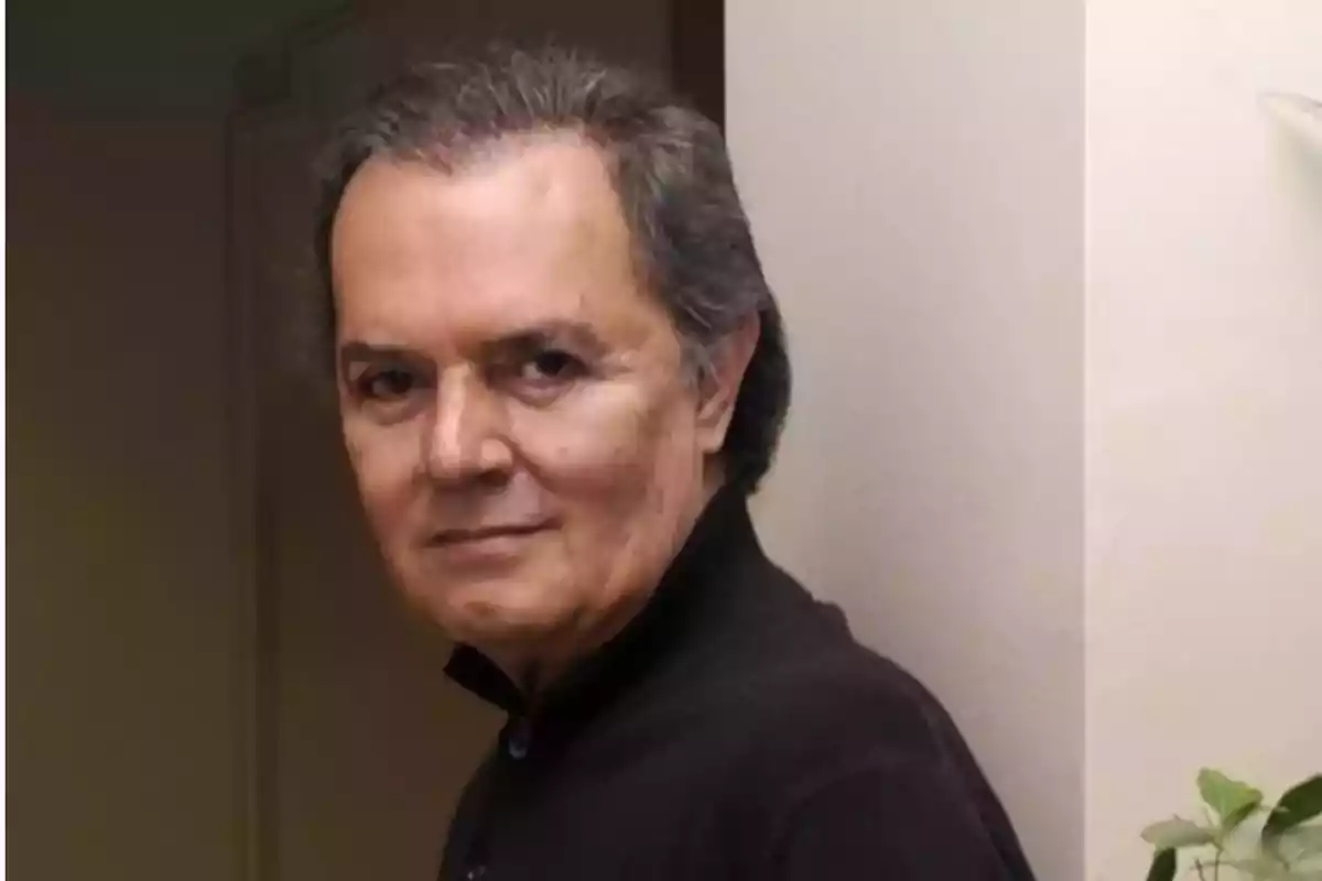 Un hombre de cabello canoso y expresión seria, vestido con una camisa oscura, está de pie junto a una pared clara. Un hombre de cabello canoso y expresión seria, vestido con una camisa oscura, está de pie junto a una pared clara.