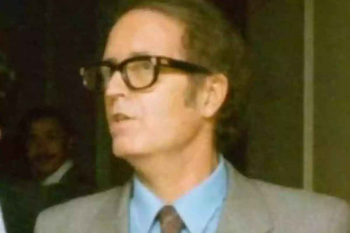 Un hombre con gafas y traje habla mientras está de pie en un entorno interior. Un hombre con gafas y traje habla mientras está de pie en un entorno interior.