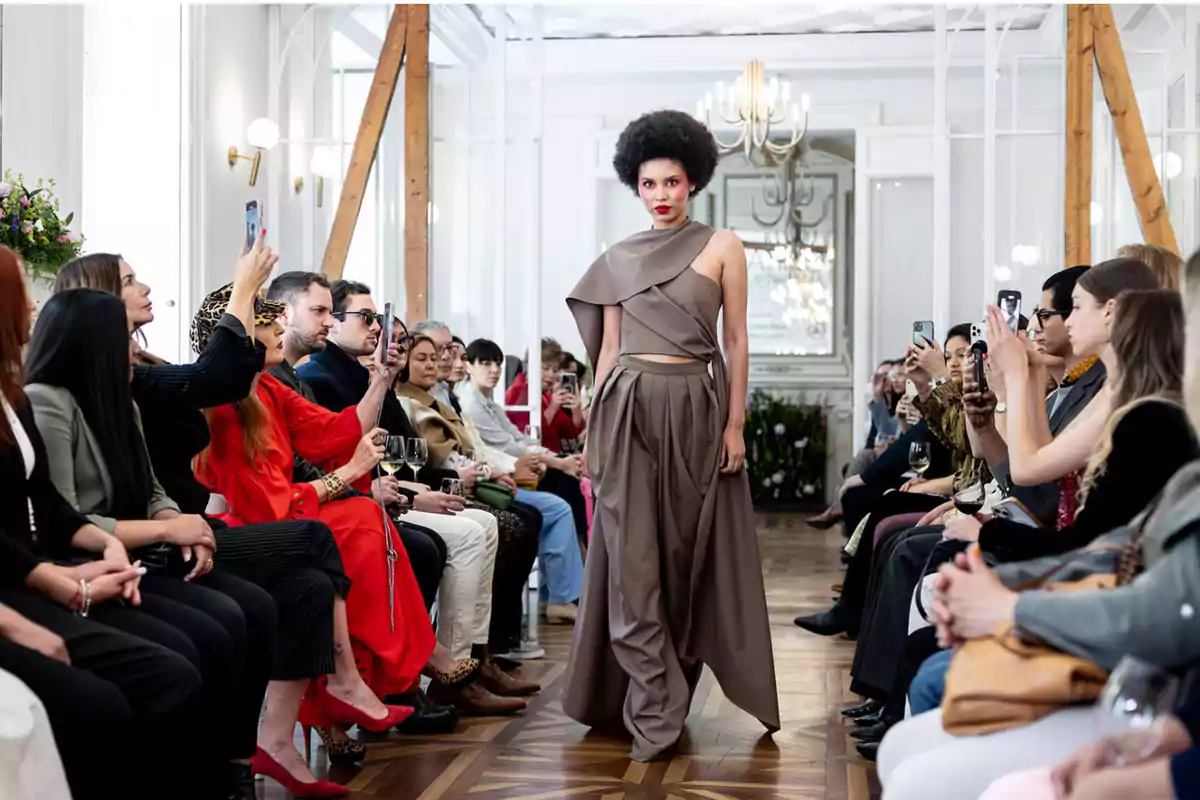Una modelo camina por la pasarela durante un desfile de moda, mientras el público observa y toma fotos con sus teléfonos móviles.