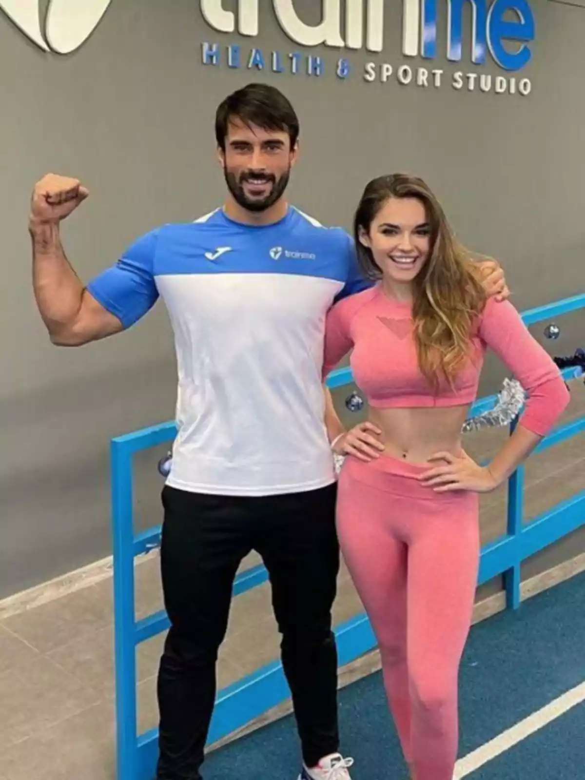 Dos personas posan en un gimnasio, una de ellas levanta el brazo mostrando el bíceps mientras la otra sonríe, ambos visten ropa deportiva. Dos personas posan en un gimnasio, una de ellas levanta el brazo mostrando el bíceps mientras la otra sonríe, ambos visten ropa deportiva.