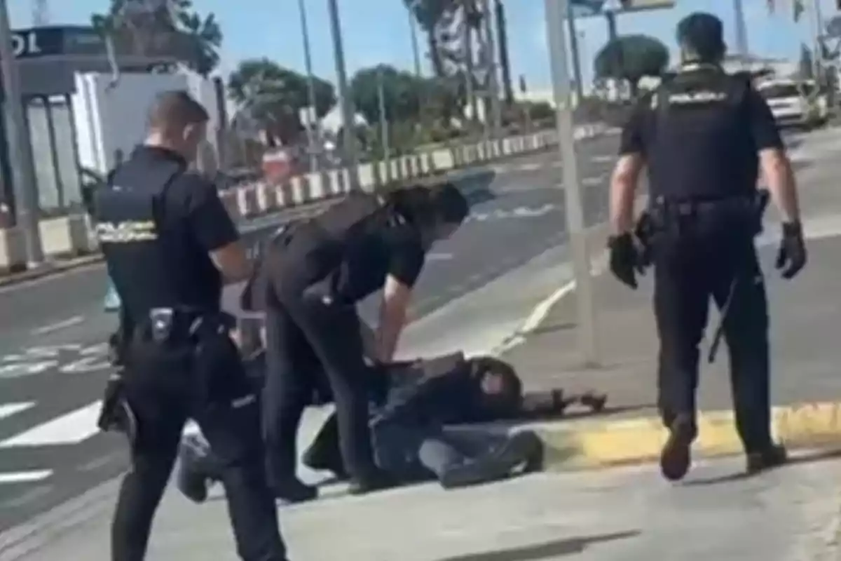 Un grupo de policías detiene a una persona en el suelo junto a una acera en una calle urbana.