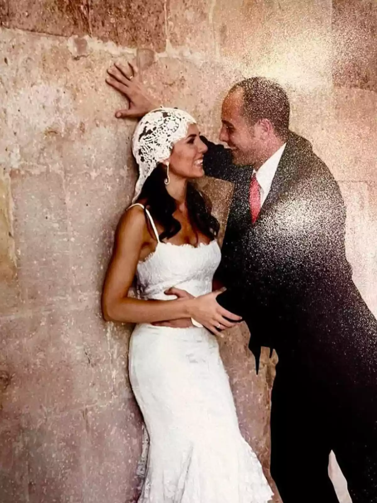 Una pareja vestida de novios se mira con cariño mientras se apoya en una pared de piedra. Una pareja vestida de novios se mira con cariño mientras se apoya en una pared de piedra.
