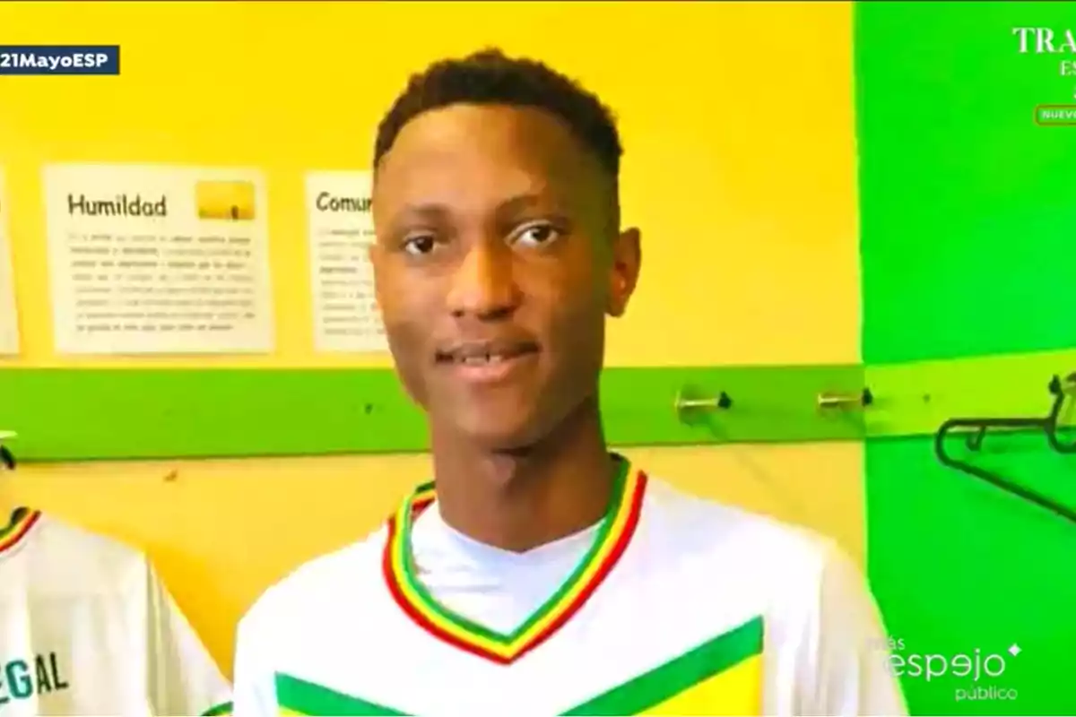 Un joven con una camiseta deportiva blanca con detalles en verde amarillo y rojo está de pie en un vestuario con paredes amarillas y verdes. Un joven con una camiseta deportiva blanca con detalles en verde amarillo y rojo está de pie en un vestuario con paredes amarillas y verdes.