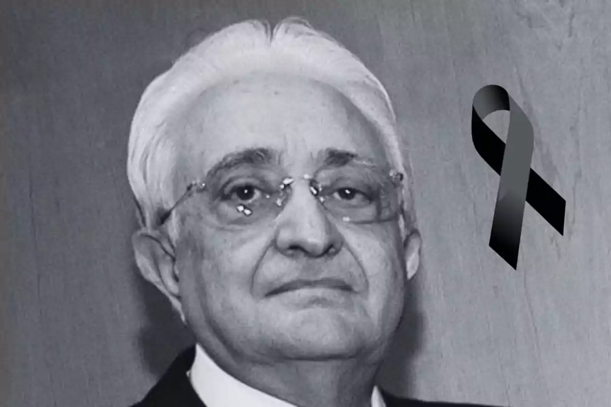 Un hombre mayor con gafas y cabello canoso aparece en una fotografía en blanco y negro con un lazo negro en señal de luto.