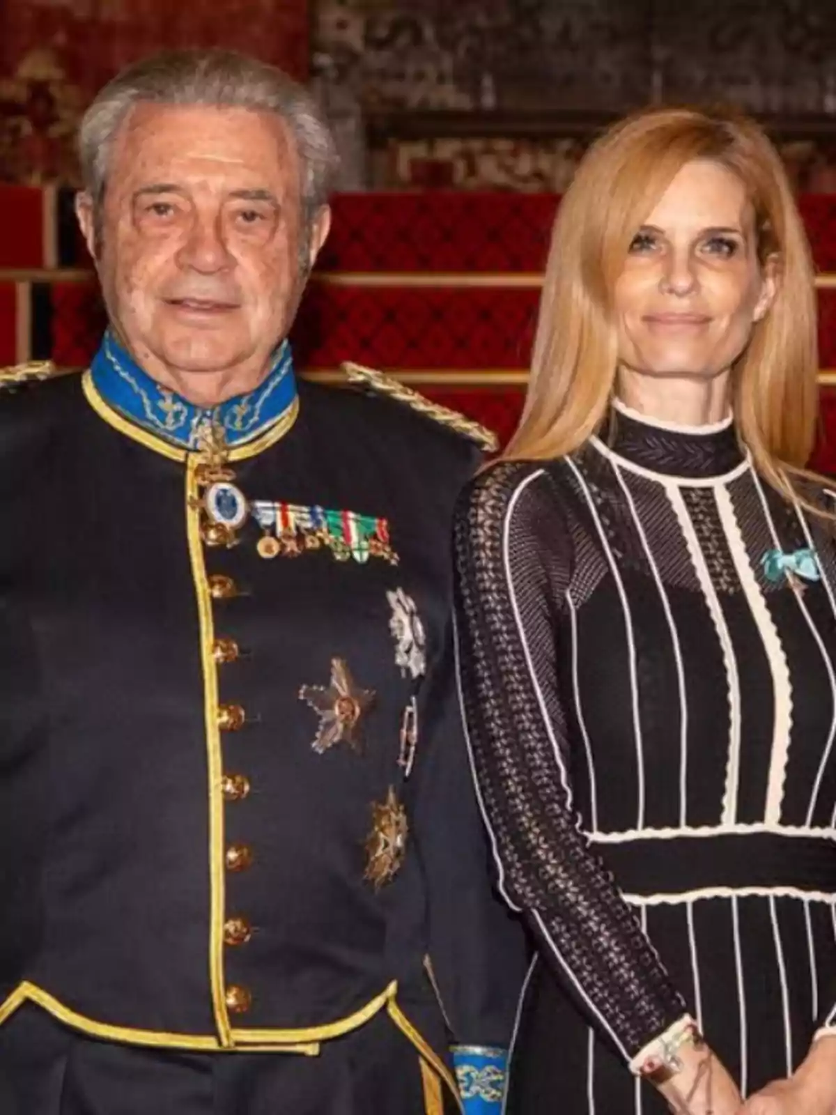 Dos personas posan juntas, una de ellas lleva un uniforme con medallas y la otra un vestido elegante, ambos están en un entorno decorado. Dos personas posan juntas, una de ellas lleva un uniforme con medallas y la otra un vestido elegante, ambos están en un entorno decorado.