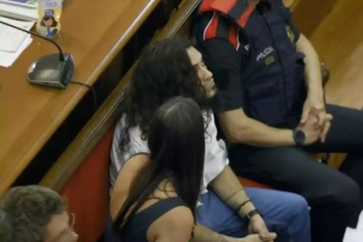 Una persona con cabello largo y rizado está sentada junto a otra persona y un oficial de policía en una sala, posiblemente en un entorno judicial. Una persona con cabello largo y rizado está sentada junto a otra persona y un oficial de policía en una sala, posiblemente en un entorno judicial.