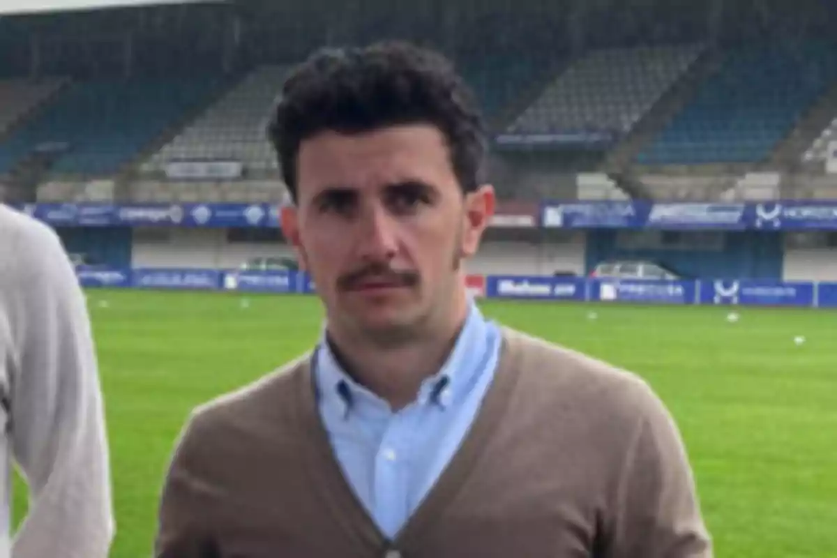 Un hombre con bigote y cabello rizado está de pie en un campo de fútbol, vistiendo una camisa azul y un suéter marrón claro. Un hombre con bigote y cabello rizado está de pie en un campo de fútbol, vistiendo una camisa azul y un suéter marrón claro.