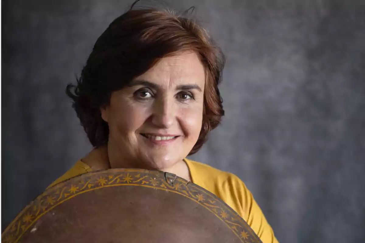 Woman smiling with brown hair and a yellow blouse holding a round object decorated with golden patterns against a gray background.