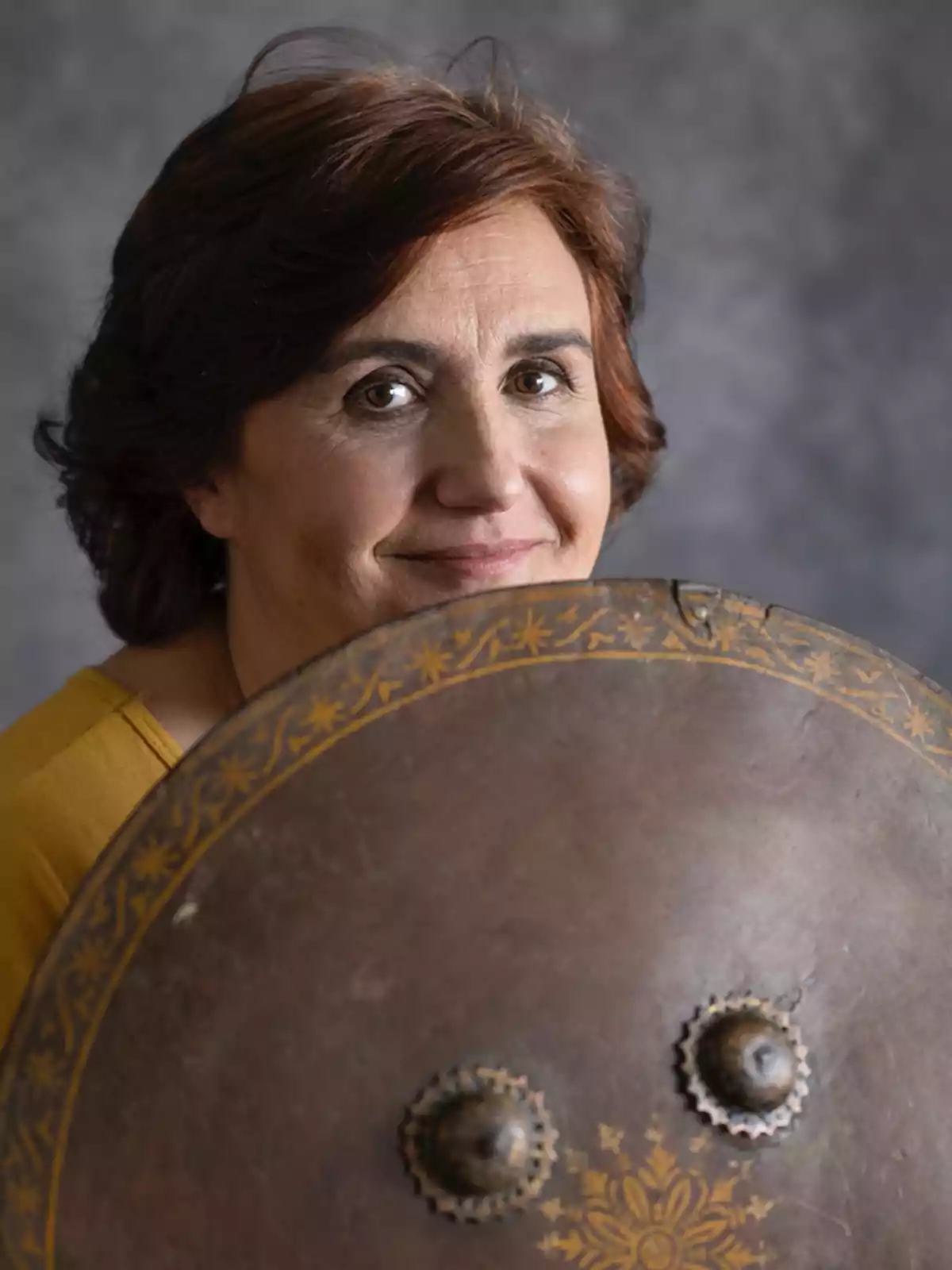 A person with brown hair and a kind expression holds a shield decorated with golden details against a gray background. A person with brown hair and a kind expression holds a shield decorated with golden details against a gray background.