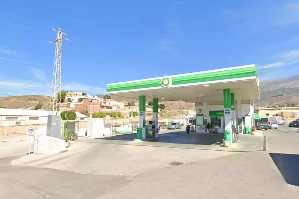 Una gasolinera con techo verde y blanco ubicada en un entorno rural con colinas y casas al fondo.