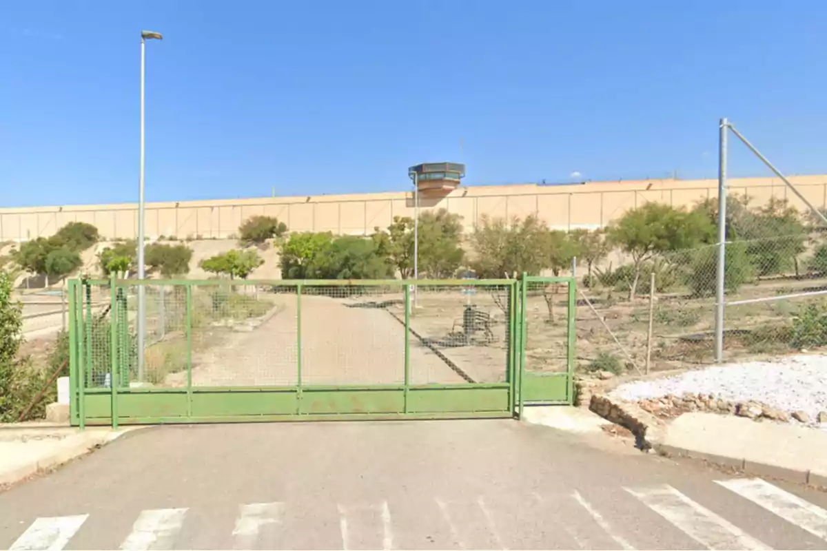 Una puerta verde de metal cerrada en un camino de acceso con una cerca y un muro alto al fondo. Una puerta verde de metal cerrada en un camino de acceso con una cerca y un muro alto al fondo.