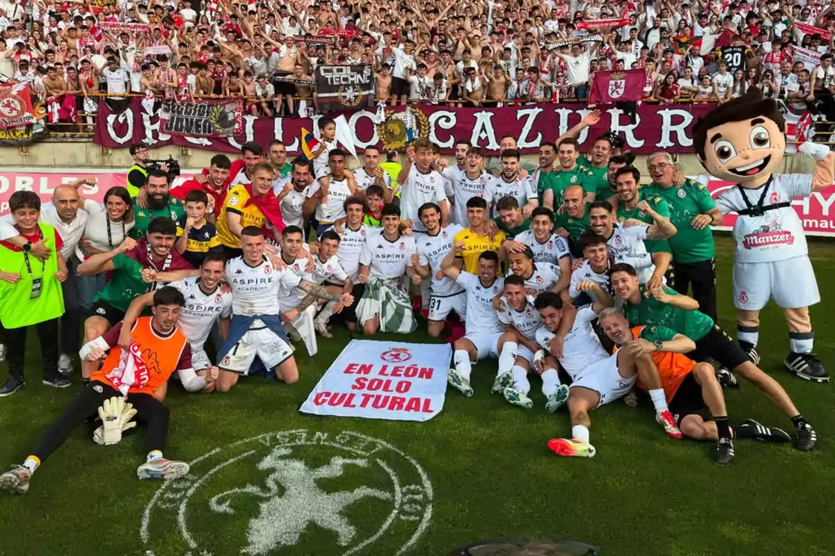Un grupo de jugadores de fútbol y aficionados celebran juntos en el campo, con una pancarta que dice "En León solo Cultural" y una mascota a la derecha.