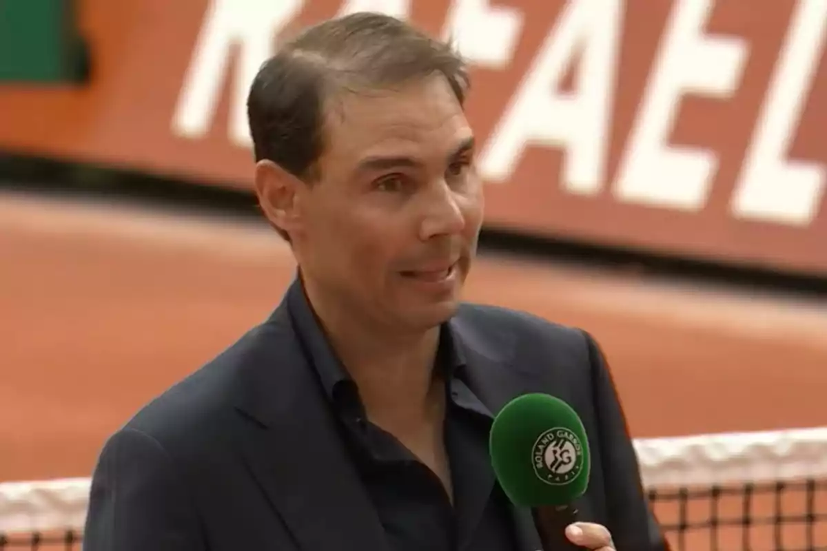 Un hombre con traje oscuro sostiene un micrófono verde en una cancha de tenis.