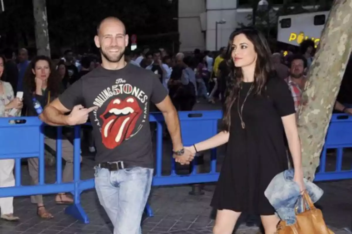 Un hombre sonriente con camiseta de los Rolling Stones sostiene la mano de una mujer vestida de negro mientras caminan juntos en un evento al aire libre rodeados de personas y vallas azules Un hombre sonriente con camiseta de los Rolling Stones sostiene la mano de una mujer vestida de negro mientras caminan juntos en un evento al aire libre rodeados de personas y vallas azules