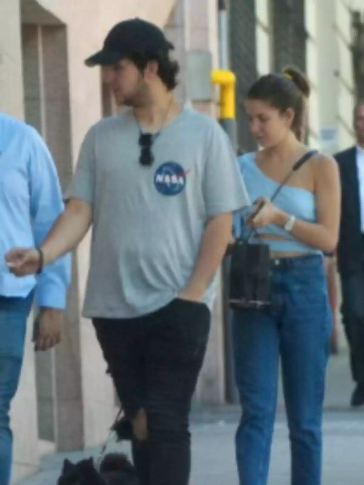 Dos personas jóvenes caminan por la calle, una de ellas lleva una camiseta gris con el logo de la NASA y gorra negra, la otra viste un top celeste y jeans, ambos parecen estar acompañados de un perro negro Dos personas jóvenes caminan por la calle, una de ellas lleva una camiseta gris con el logo de la NASA y gorra negra, la otra viste un top celeste y jeans, ambos parecen estar acompañados de un perro negro