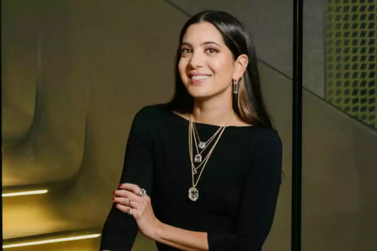 Mujer joven de cabello largo y oscuro con vestido negro y joyería elegante sonriendo frente a una escalera moderna